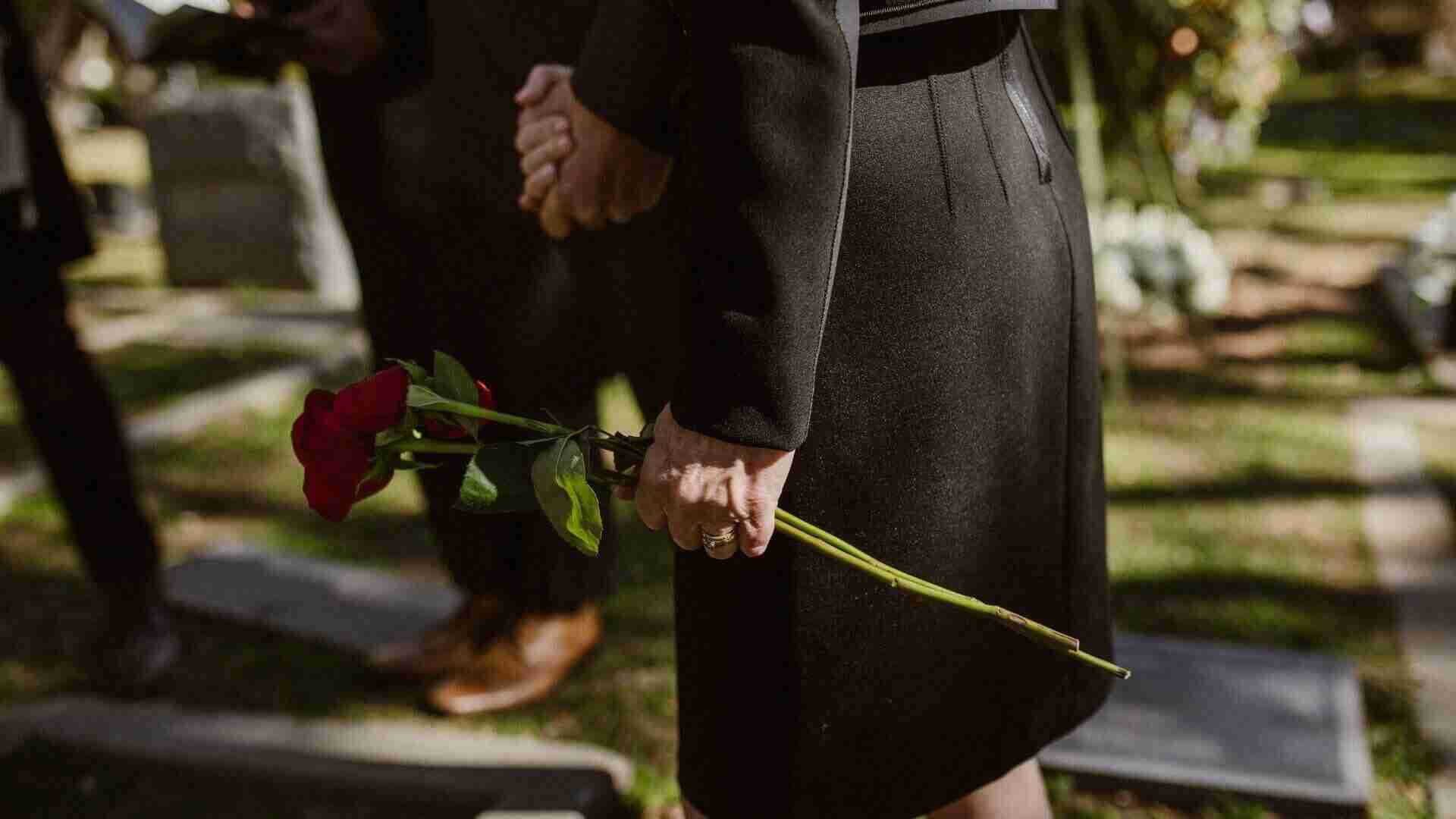 Frau in schwarzer Kleidung hält eine rote Rose in der Hand auf einem Friedhof.