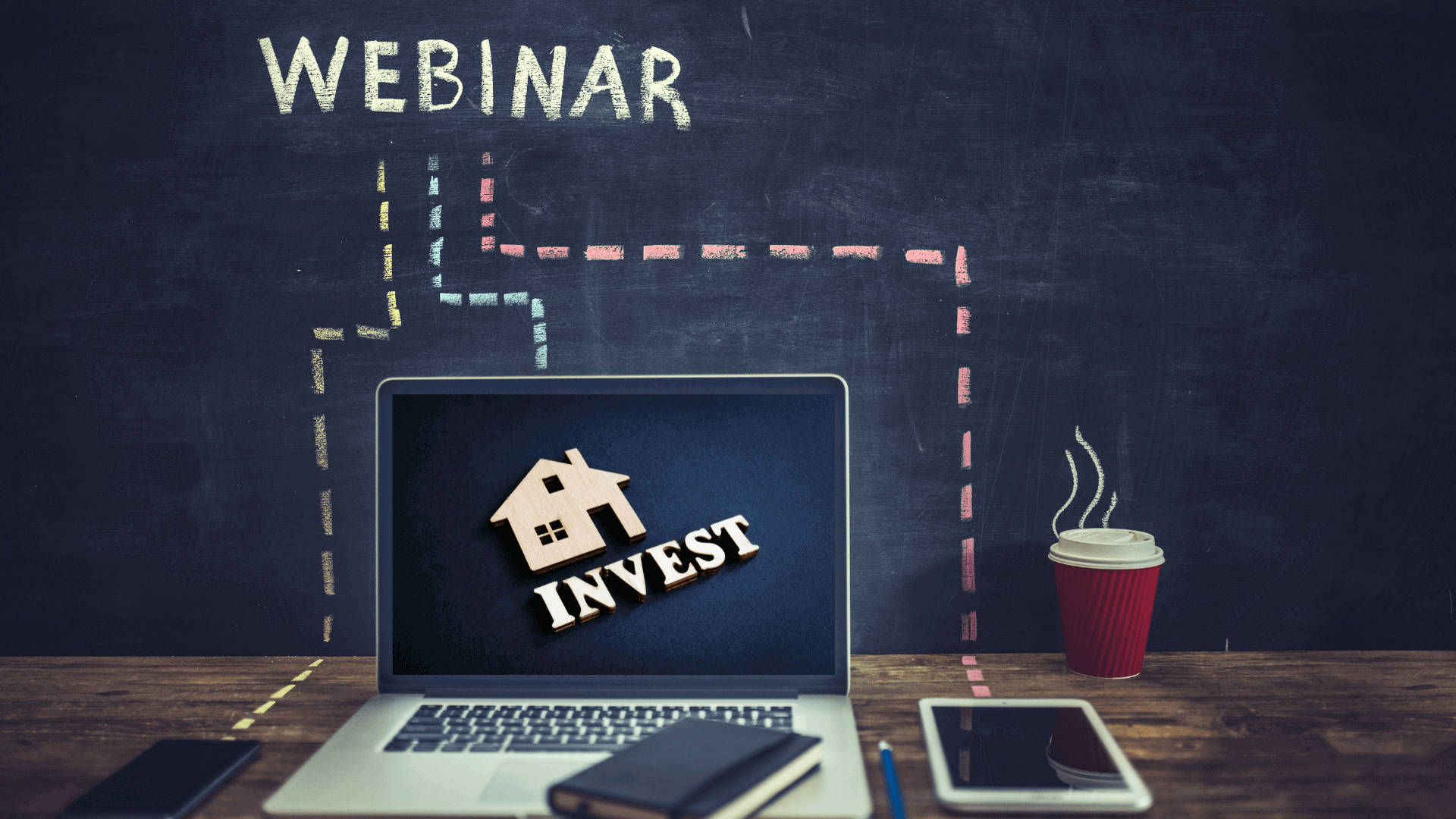Laptop mit dem Wort 'INVEST' und einem Haus-Symbol auf dem Bildschirm, daneben ein Notizbuch und ein Getränk.